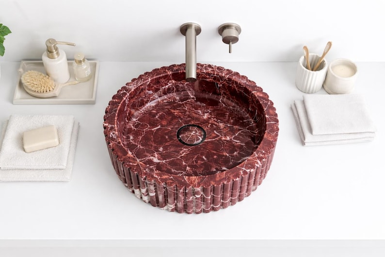 Peut inclure: Un lavabo rond en marbre bordeaux avec un ext&eacute;rieur cannel&eacute;. Le lavabo est pos&eacute; sur une surface blanche, avec un robinet argent&eacute; et d'autres accessoires de salle de bain, dont un distributeur de savon, du savon et des serviettes. L'int&eacute;rieur du lavabo pr&eacute;sente un motif marbr&eacute;.