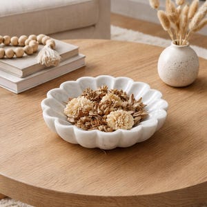 May include: A white marble decorative bowl with a scalloped edge, filled with dried flowers. The bowl sits on a round wooden coffee table. A small vase with dried plants and a stack of books are also on the table.