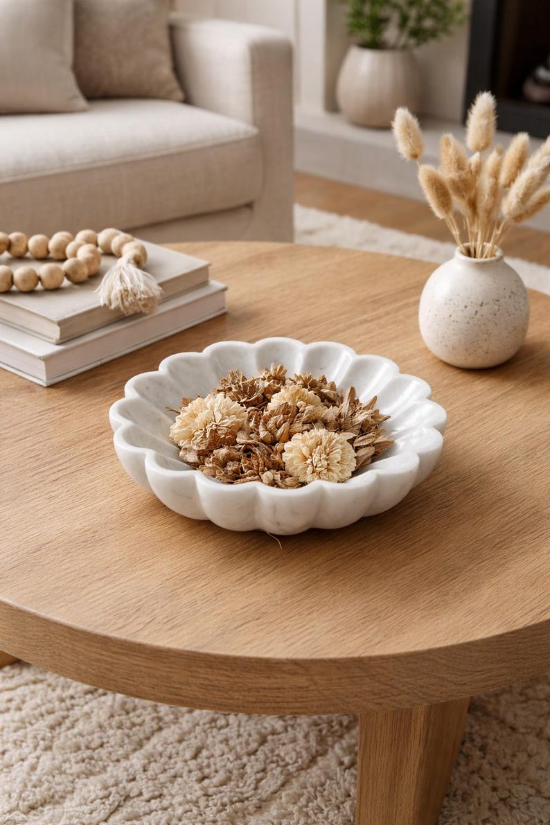 May include: A white marble decorative bowl with a scalloped edge, filled with dried flowers. The bowl sits on a round wooden coffee table. A small vase with dried plants and a stack of books are also on the table.