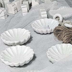 May include: A collection of white marble decorative bowls with a scalloped edge design. The bowls are arranged on a textured surface, alongside other marble pieces and a bundle of twine.