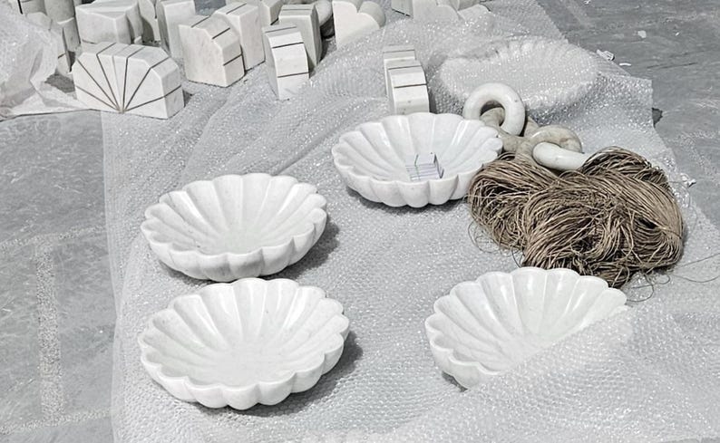 May include: A collection of white marble decorative bowls with a scalloped edge design. The bowls are arranged on a textured surface, alongside other marble pieces and a bundle of twine.
