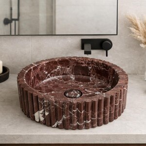 Peut inclure: Un lavabo rond en marbre rouge avec un ext&eacute;rieur cannel&eacute;. Le lavabo est pos&eacute; sur un comptoir de couleur claire dans une salle de bain. Autres accessoires de salle de bain : porte-savon, distributeur de savon et vase avec fleurs s&eacute;ch&eacute;es.
