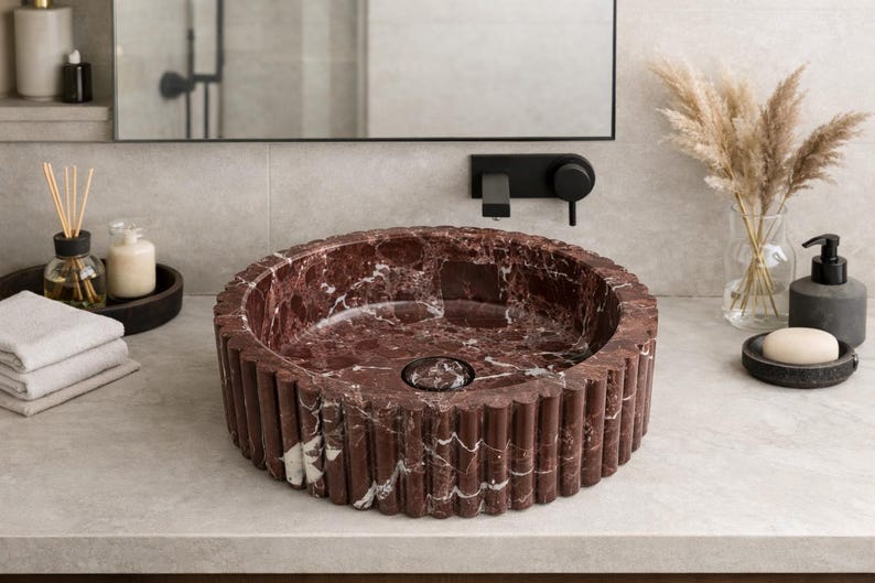 Peut inclure: Un lavabo rond en marbre rouge avec un ext&eacute;rieur cannel&eacute;. Le lavabo est pos&eacute; sur un comptoir de couleur claire dans une salle de bain. Autres accessoires de salle de bain : porte-savon, distributeur de savon et vase avec fleurs s&eacute;ch&eacute;es.