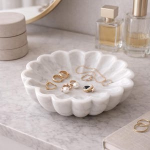 May include: A white marble jewelry dish with a scalloped edge holds various gold rings, earrings, and a necklace. Two perfume bottles and a small stack of boxes are in the background. The dish sits on a marble surface.