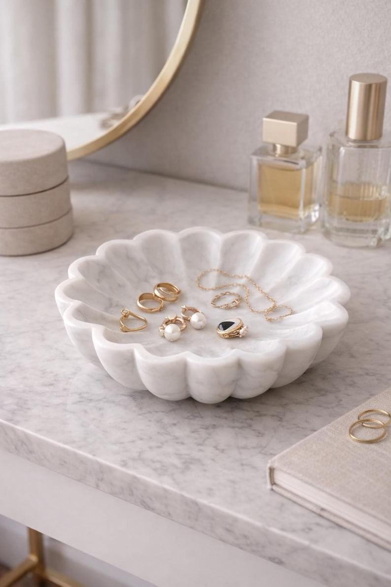 May include: A white marble jewelry dish with a scalloped edge holds various gold rings, earrings, and a necklace. Two perfume bottles and a small stack of boxes are in the background. The dish sits on a marble surface.