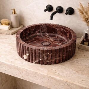 Peut inclure: Un lavabo de salle de bain rond en marbre rouge avec un ext&eacute;rieur cannel&eacute;. L'&eacute;vier pr&eacute;sente un motif vein&eacute; rouge fonc&eacute; et blanc. Un robinet et des accessoires noirs sont mont&eacute;s sur le mur au-dessus de l'&eacute;vier. L'&eacute;vier est sur un comptoir clair.