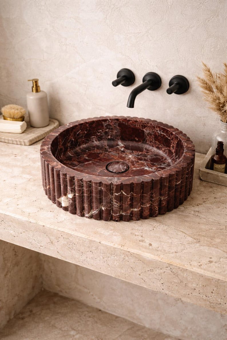 Peut inclure: Un lavabo de salle de bain rond en marbre rouge avec un ext&eacute;rieur cannel&eacute;. L'&eacute;vier pr&eacute;sente un motif vein&eacute; rouge fonc&eacute; et blanc. Un robinet et des accessoires noirs sont mont&eacute;s sur le mur au-dessus de l'&eacute;vier. L'&eacute;vier est sur un comptoir clair.