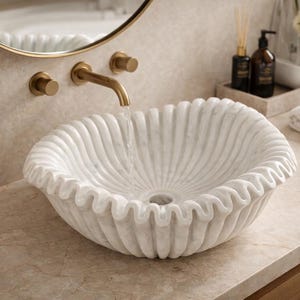 May include: A white marble sink with a scalloped design, resembling a seashell. The sink is set on a light-colored countertop, with a gold faucet and a round mirror in the background. The water is running from the faucet into the sink.