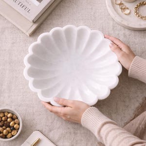 May include: A white marble decorative bowl with a scalloped edge. The bowl is held by two hands. The bowl is on a beige surface, with a small bowl of nuts, a stack of books, and a candle on a tray in the background.
