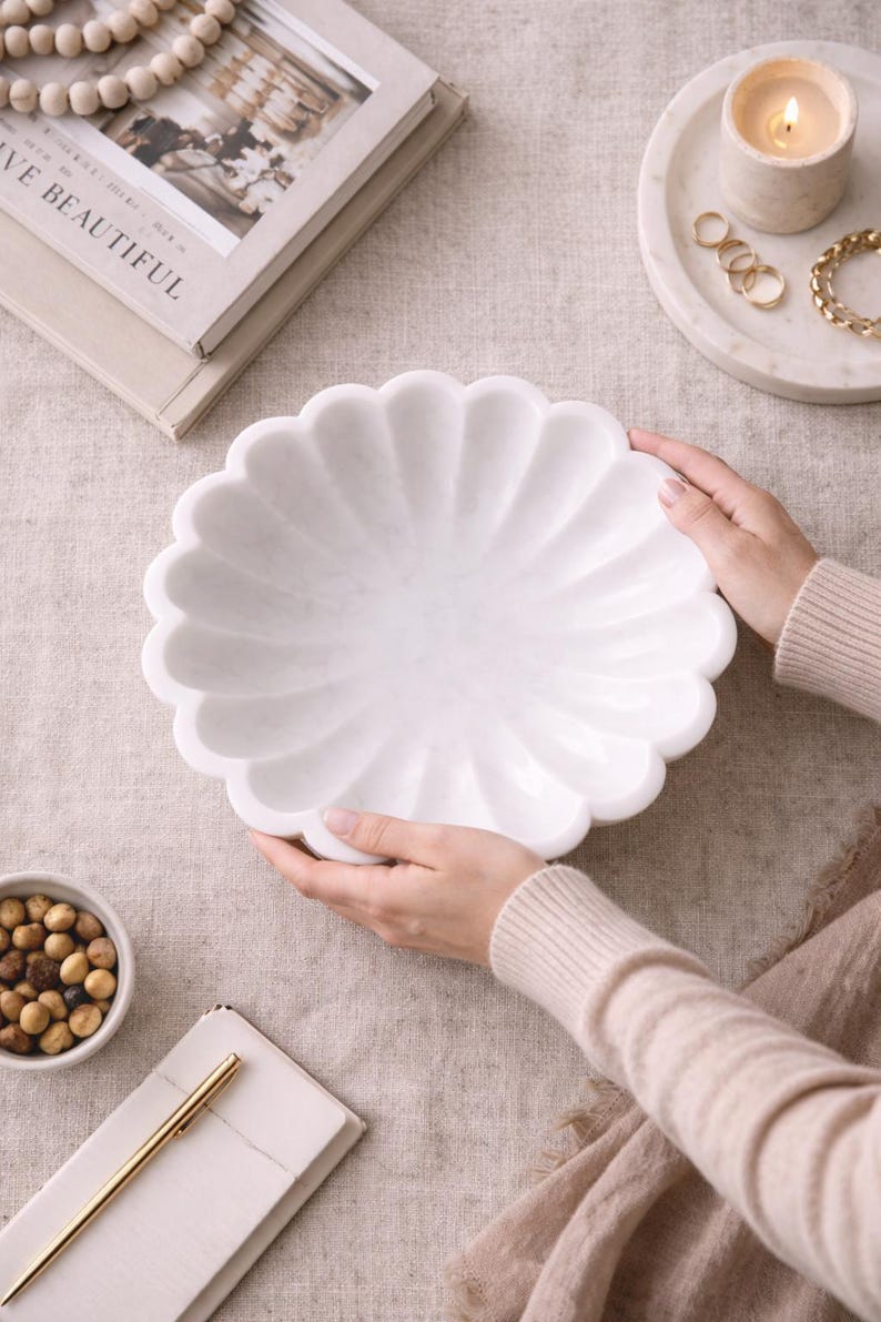 May include: A white marble decorative bowl with a scalloped edge. The bowl is held by two hands. The bowl is on a beige surface, with a small bowl of nuts, a stack of books, and a candle on a tray in the background.