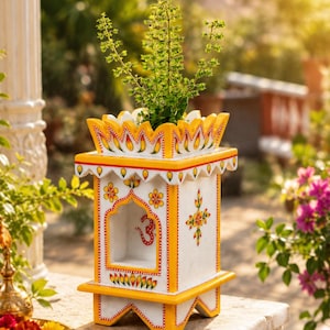 May include: A white and yellow painted wooden planter with a green plant. The planter has a decorative design with floral and geometric patterns. The planter is sitting on a stone surface, with a blurred background of greenery and flowers.