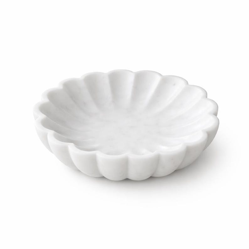 May include: A white marble decorative bowl with a scalloped edge. The bowl is round and has a shallow interior. The marble has a smooth, polished finish. The bowl is suitable for holding small items.