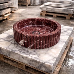 Peut inclure: Un &eacute;vier rond en marbre rouge avec un ext&eacute;rieur cannel&eacute;, pos&eacute; sur une grande dalle de marbre. L'&eacute;vier a un drain sombre et repose sur une palette en bois. Le marbre pr&eacute;sente des veines blanches.