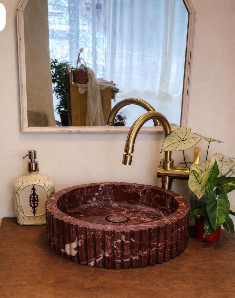 Peut inclure: Un &eacute;vier en marbre rouge avec un ext&eacute;rieur cannel&eacute; repose sur une surface en bois. Un robinet dor&eacute; et un distributeur de savon d&eacute;coratif sont visibles. Un miroir et de la verdure sont en arri&egrave;re-plan.