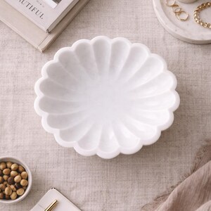 May include: A white marble decorative bowl with a scalloped edge. The bowl is on a neutral-colored linen surface, alongside a small bowl of nuts, a candle, and a stack of books. Other decorative items include a gold pen, rings, and a bracelet.