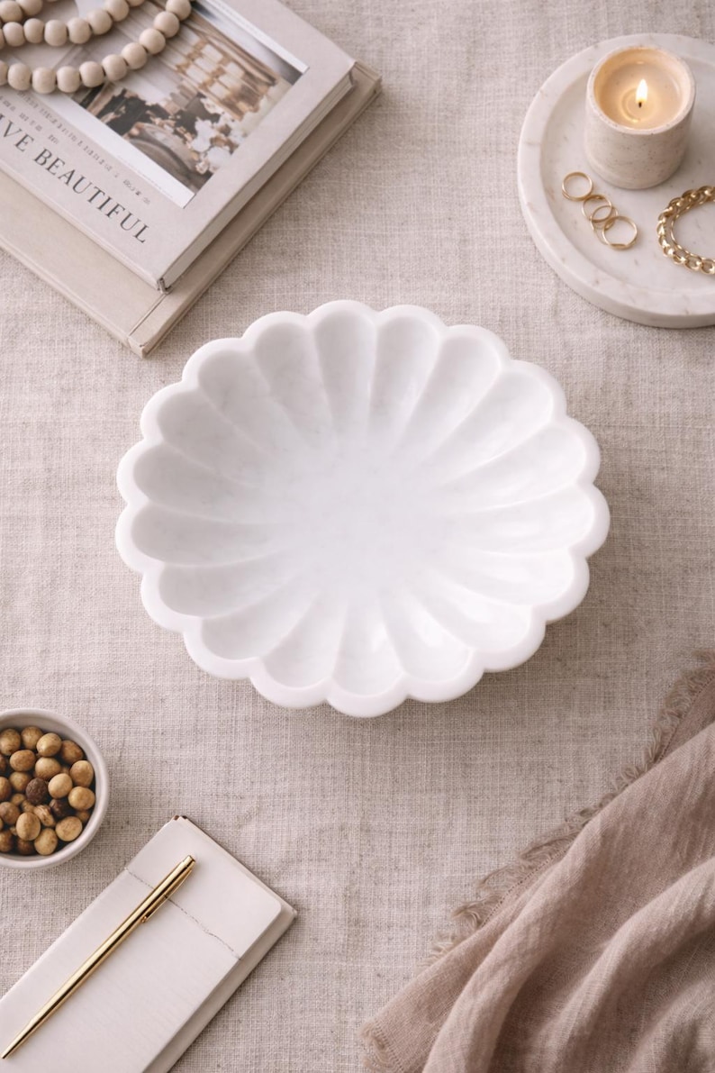 May include: A white marble decorative bowl with a scalloped edge. The bowl is on a neutral-colored linen surface, alongside a small bowl of nuts, a candle, and a stack of books. Other decorative items include a gold pen, rings, and a bracelet.