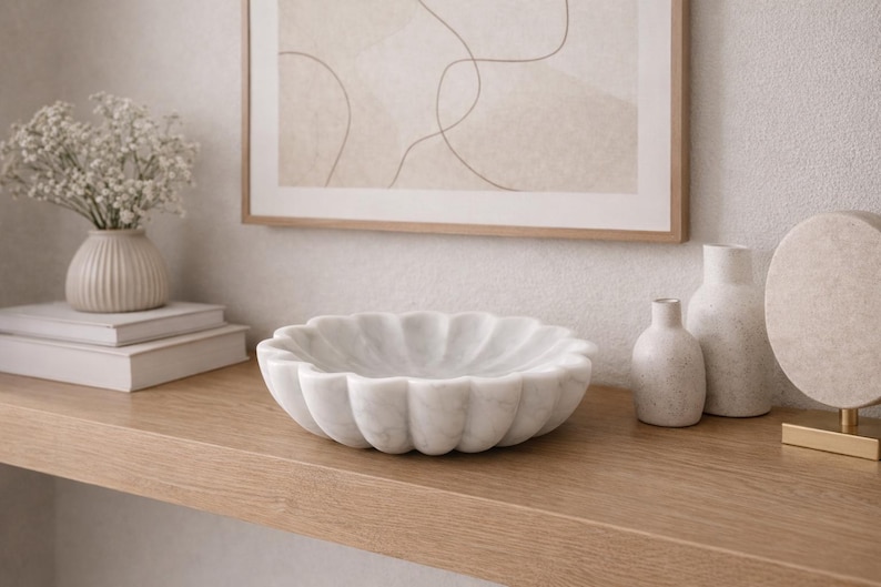 May include: A white marble bowl with a scalloped edge sits on a wooden shelf. Beside it are two small vases, a larger vase, and a decorative round object. A framed artwork hangs on the wall above the shelf, and a small vase of flowers sits on a stack of books.