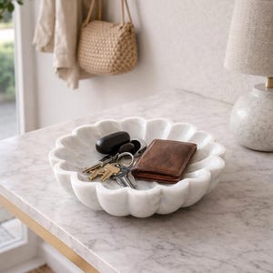 May include: A white marble decorative bowl with a scalloped edge, holding keys and a brown leather wallet. The bowl sits on a marble-topped table with a gold-colored metal frame. A beige handbag hangs in the background.