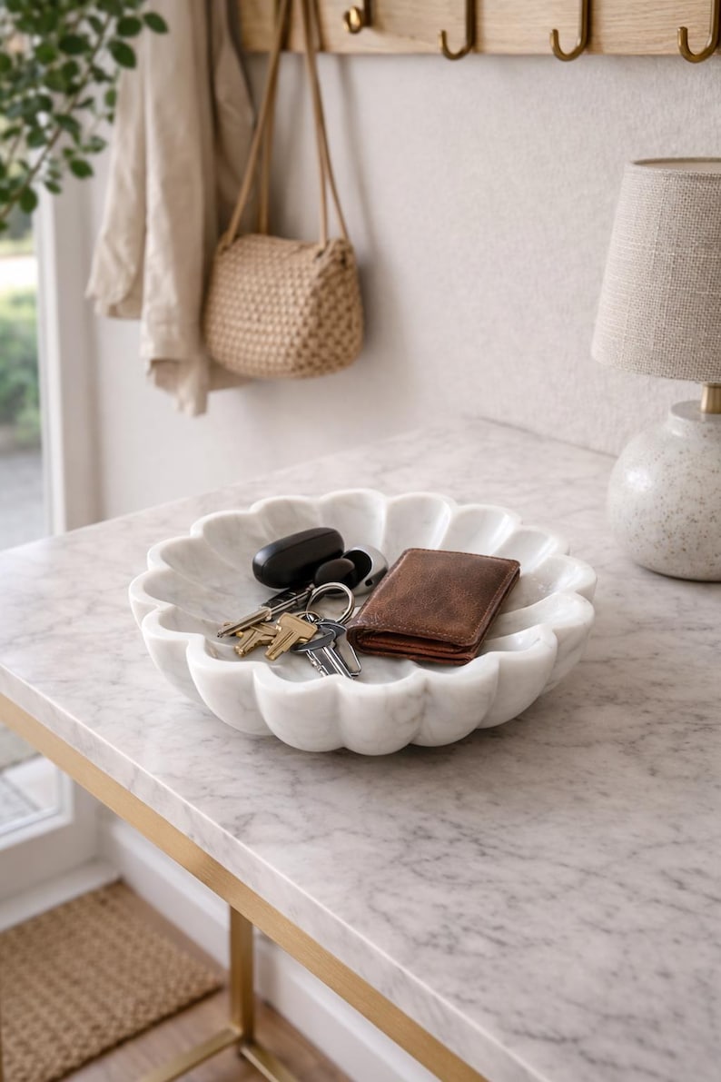 May include: A white marble decorative bowl with a scalloped edge, holding keys and a brown leather wallet. The bowl sits on a marble-topped table with a gold-colored metal frame. A beige handbag hangs in the background.
