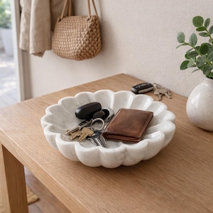 May include: A white marble decorative bowl with a scalloped edge holds keys and a brown leather wallet. The bowl sits on a light wood table, with a small vase and a woven bag in the background.