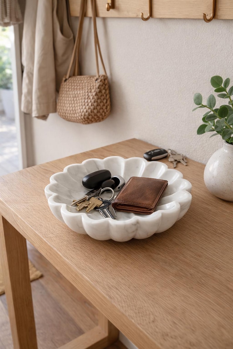 May include: A white marble decorative bowl with a scalloped edge holds keys and a brown leather wallet. The bowl sits on a light wood table, with a small vase and a woven bag in the background.