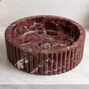 Peut inclure: Un lavabo rond en marbre bordeaux avec un ext&eacute;rieur cannel&eacute;. Le lavabo a un drain central et est pos&eacute; sur un comptoir blanc. C'est un &eacute;l&eacute;ment de salle de bain.