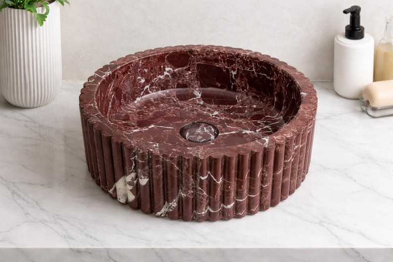 Peut inclure: Un lavabo rond en marbre bordeaux avec un ext&eacute;rieur cannel&eacute;. Le lavabo a un drain central et est pos&eacute; sur un comptoir blanc. C'est un &eacute;l&eacute;ment de salle de bain.
