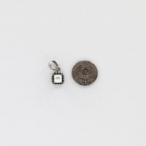 May include: A small, square pendant with a pearl center and a silver-colored, beaded frame. The pendant is shown next to a quarter for size comparison. The pendant has a small loop at the top for a chain.