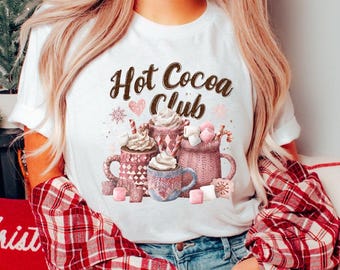 May include: White t-shirt with the text "Hot Cocoa Club" in brown script. The shirt features a graphic of several mugs of hot cocoa with whipped cream, marshmallows, and decorative elements. The person is wearing a red and white plaid shirt.