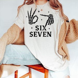 May include: Cream-colored t-shirt featuring a black skeleton hand design making the 'okay' gesture and pointing. The text "SIX SEVEN" is printed below the hands in a bold, black font. Small star accents are placed around the hands and text.