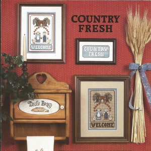 May include: A collection of country-themed cross-stitch art and decor. Includes framed pieces with "WELCOME" and "COUNTRY FRESH" text, a wooden bread box, a towel with a chicken design, and a bundle of wheat tied with a blue ribbon. The background is a red textured wall.