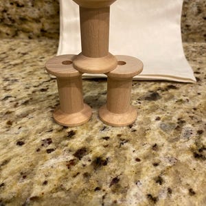 May include: Three light-colored wooden spools arranged in a pyramid formation. Each spool features a cylindrical body with a wider circular top and base, and a small hole centered through the middle of each piece, showcasing a smooth, natural wood grain.