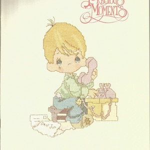 May include: Cross-stitch pattern featuring a blonde-haired child holding a pink telephone, seated at a desk. The image includes the text "Precious Moments" and "Dear Jon". The color palette includes pastel shades of pink, blue, yellow, and green.
