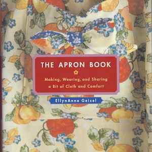 May include: A book titled "The Apron Book" with a cover featuring a yellow apron with a fruit and floral pattern. The apron has a bow detail and the book's title is in a red box. The text reads "Making, Wearing, and Sharing a Bit of Cloth and Comfort."