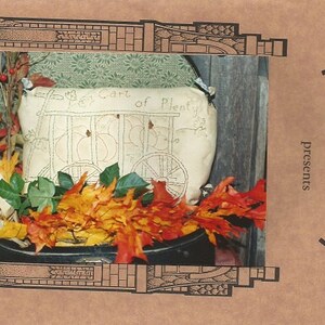 May include: A beige pillow with a stitched design of a cart filled with pumpkins, labeled "Cart of Plenty." The pillow is surrounded by fall foliage, including orange and yellow leaves. The image is framed with a brown border and the text "Mom & / presents" is visible.