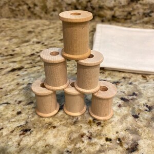 May include: A stack of six light-colored wooden spools arranged in a pyramid formation. Each spool features a cylindrical body with a central hole and flared top and bottom edges, showcasing a natural wood grain texture and a smooth, unfinished surface.