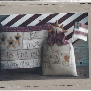 May include: A patriotic decorative piece featuring a pillow and a figure holding a small flag. The pillow displays a flag design with stars and the text "Be true to the red, white and blue." The figure has a star-shaped pocket.