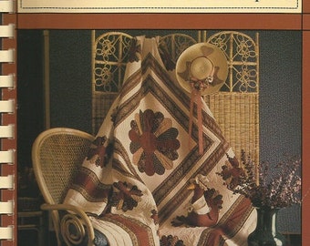 May include: The cover of a book titled "The Pieceable Kingdom" by Mary Jane Lamphier. The image features a quilt with a floral design in shades of cream, brown, and burgundy, draped over a wicker chair. A straw hat with a ribbon hangs nearby.