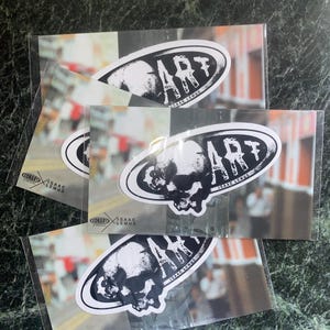 Könnte beinhalten: Mehrere Aufkleber mit einem schwarz-weißen Schädeldesign und dem Wort "ART" in fetten Buchstaben. Die Aufkleber befinden sich in einer transparenten Kunststoffverpackung, mit dem Namen "ISAAC LEMUS" darunter. Der Hintergrund ist eine verschwommene Straßenszene.