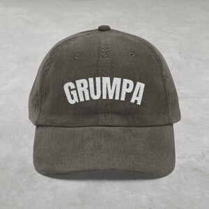 May include: Olive green corduroy baseball cap with the word "GRUMPA" embroidered in white block letters across the front. The cap has a curved brim and a small button on top.