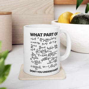 Könnte beinhalten: Weiße Keramik-Tasse mit dem Text "WHAT PART OF DON'T YOU UNDERSTAND?" und mathematischen Gleichungen in Schwarz. Die Tasse steht auf einem beigen Untersetzer.