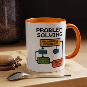 Może przedstawiać: Biały ceramiczny kubek z pomarańczowym wnętrzem i uchwytem. Kubek zawiera napis "PROBLEM SOLVING" i schemat blokowy z pytaniami "DO YOU HAVE A PROBLEM?", "CAN YOU SOLVE IT?" i "DON'T WORRY". Łyżka i makaronik leżą na drewnianej powierzchni.