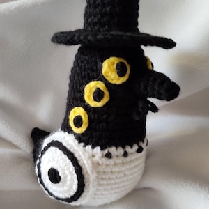 May include: A handmade crochet figure of a plague doctor, featuring a black hat and mask, with yellow eye accents. The body is black and white, with a white mask-like area. The figure is resting on a white surface.