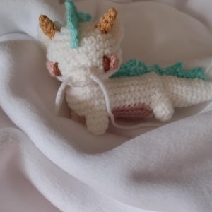 K&ouml;nnte beinhalten: Ein handgefertigter wei&szlig;er H&auml;kchendrache mit hellblauen und hellbraunen Akzenten. Der Drache hat einen langen K&ouml;rper, kleine Beine und ein s&uuml;&szlig;es Gesicht mit rosa Wangen. Der Drache liegt auf einer wei&szlig;en, weichen Oberfl&auml;che.