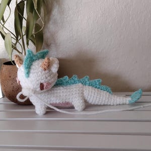 K&ouml;nnte beinhalten: Handgeh&auml;kelter Drache in Wei&szlig; mit hellblauen Stacheln und Akzenten. Der Drache hat kleine, hellbraune H&ouml;rner und rosa Wangen. Er liegt auf einer wei&szlig;en Oberfl&auml;che, im Hintergrund eine Pflanze in einem braunen Topf.