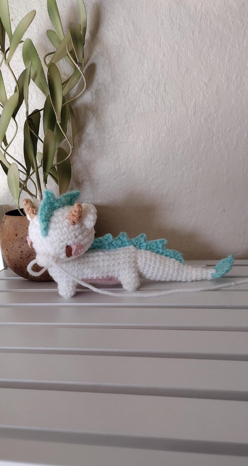 K&ouml;nnte beinhalten: Handgeh&auml;kelter Drache in Wei&szlig; mit hellblauen Stacheln und Akzenten. Der Drache hat kleine, hellbraune H&ouml;rner und rosa Wangen. Er liegt auf einer wei&szlig;en Oberfl&auml;che, im Hintergrund eine Pflanze in einem braunen Topf.
