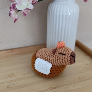 May include: A handmade crochet capybara figurine in a brown and orange color scheme, with a white patch. The capybara is resting in a crocheted nest. A white vase with purple and white orchids is in the background.