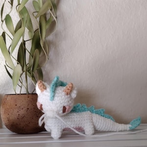 K&ouml;nnte beinhalten: Ein handgefertigter wei&szlig;er H&auml;keldrache mit hellblauen und beigen Akzenten. Der Drache hat einen langen Schwanz und liegt auf einem Regal. Der Drache ist ein niedliches, skurriles Dekorationsst&uuml;ck.