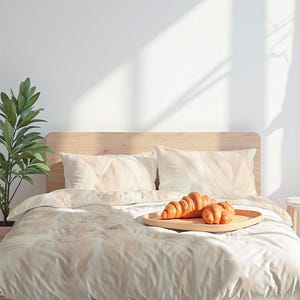 May include: A light wood bed frame with matching headboard and bedding. The bedding has a light beige pattern. Two croissants rest on a wooden tray on top of the bed. A green plant is to the left of the bed.