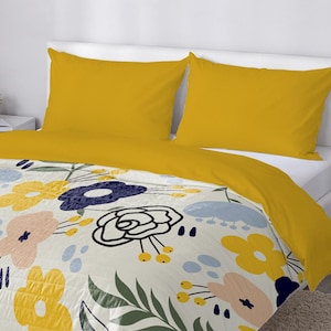 May include: A bed with a floral patterned comforter and two yellow pillowcases. The comforter features a cream background with large yellow, blue, and pink flowers. The bed is made up with a yellow top sheet.